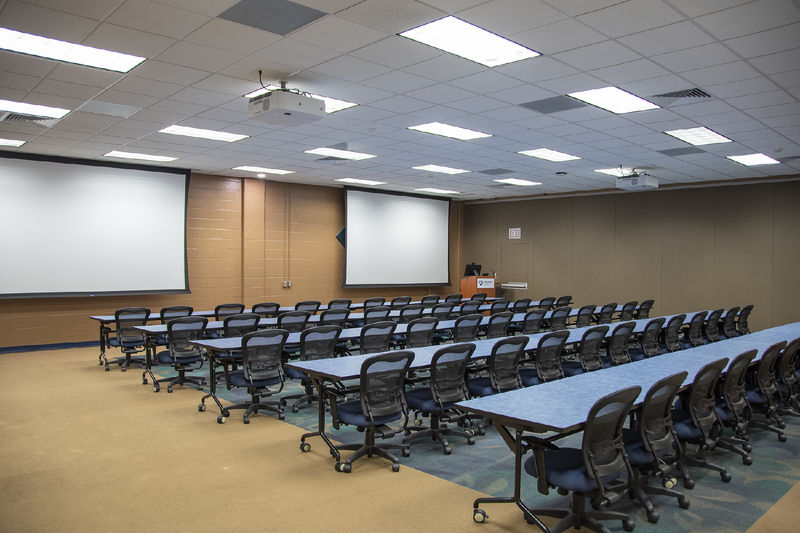 Room with brown walls and two projector screens hanging from drop ceiling with long blue tables and black chairs on wheels